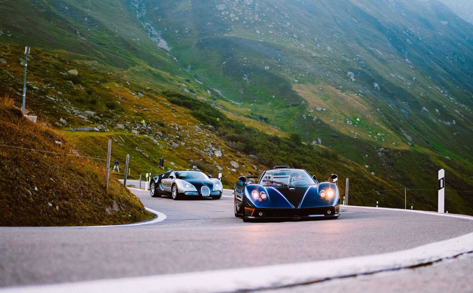 Stunning Pegani and Bugatti super cars driving along a mountain road.